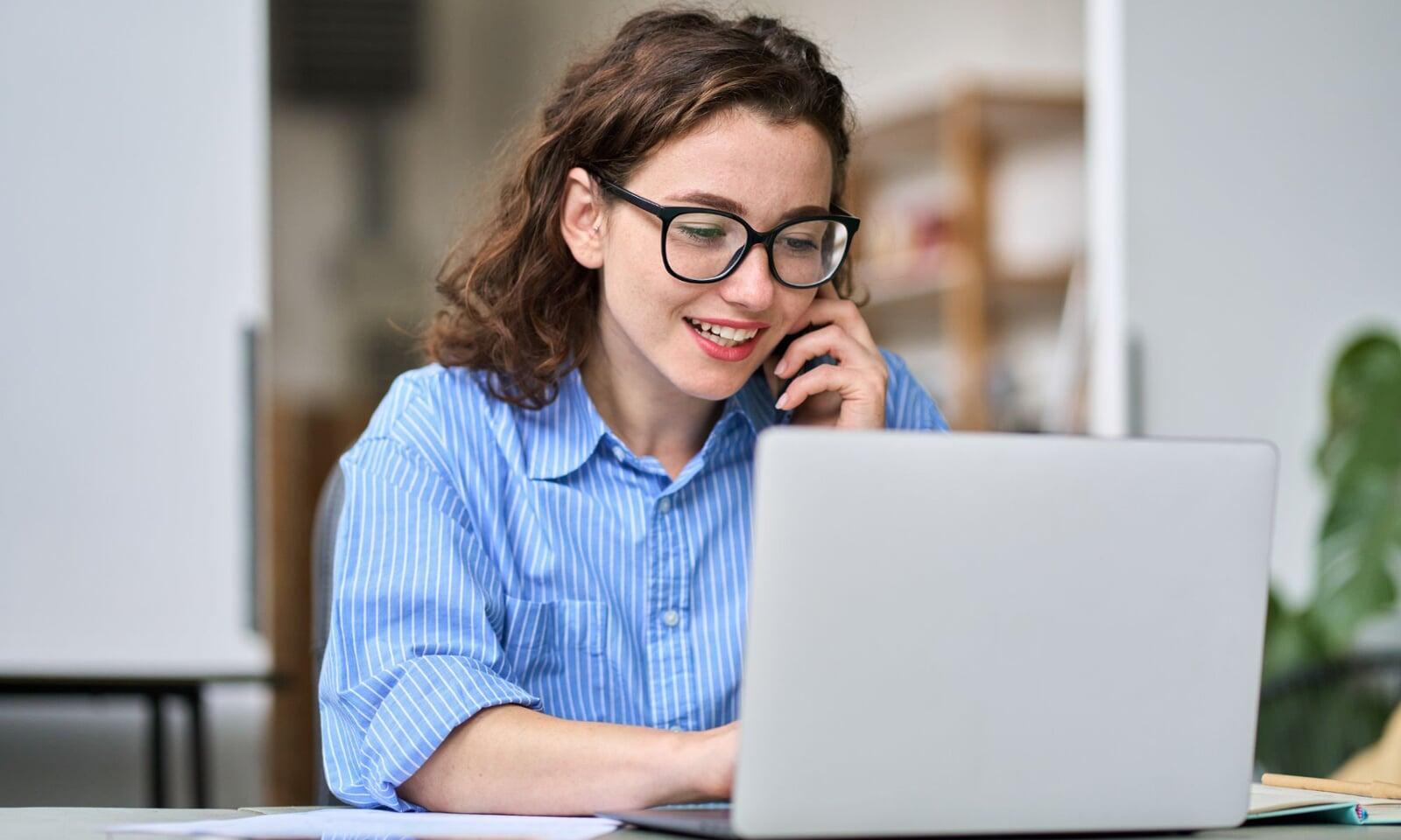 Person wearing glasses, smiling while talking on the phone and working on a laptop at a desk in a bright, modern workspace.